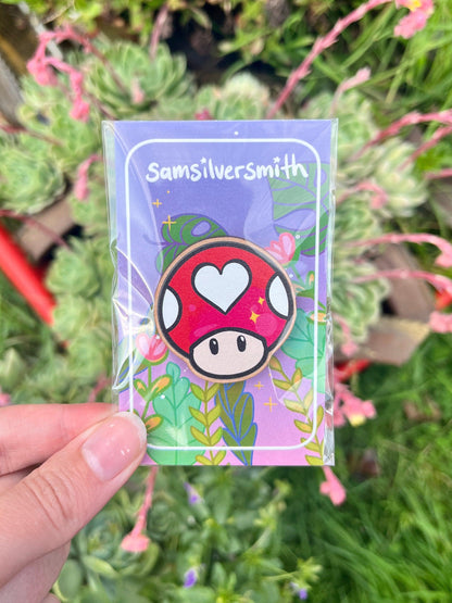 Close-up of a hand holding the Super Mushroom Wooden Pin, an artist-designed wooden accessory featuring a red mushroom with a heart on its cap. Perfect as a gift, the pin is displayed on a card labeled Samsilversmith, set against a blurred background of green foliage and pink flowers.