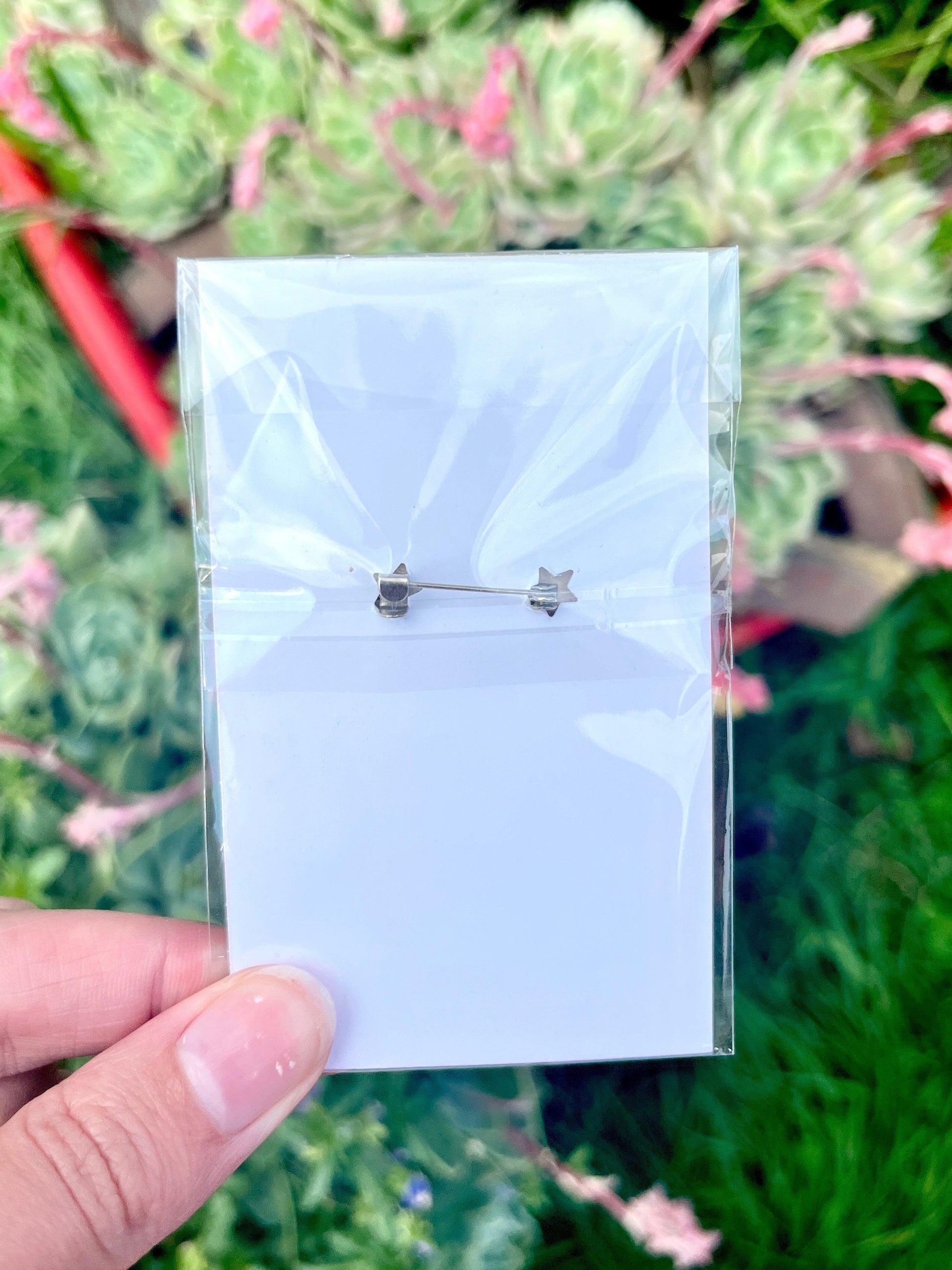 A person holds a clear plastic bag containing a Pixel Heart Wooden Pin set against a backdrop of green and pink plants.