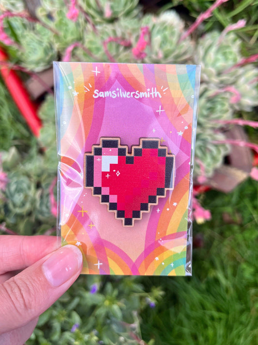 A hand holds a plastic-wrapped, artist-designed wooden pin featuring a pixelated heart. The pin is attached to a colorful card with "Samsilversmith" on it, set against a backdrop of vibrant plants and greenery.