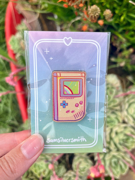 A hand displays the Game Boy Wooden Pin, designed to resemble a retro handheld gaming console. The pin, crafted from wood and featuring a colorful screen, is mounted on a card decorated with hearts and branded with the name Samsilversmith. In the background, lush green plants contrast beautifully with vibrant red accents.