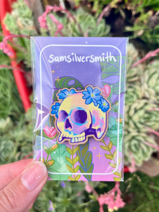 A vibrant, artist-crafted wooden pin depicting a skull adorned with flowers is showcased on a floral-themed card. The card, featuring the name Samsilversmith, is held by hand and sits against greenery that enhances its lively, nature-inspired design.