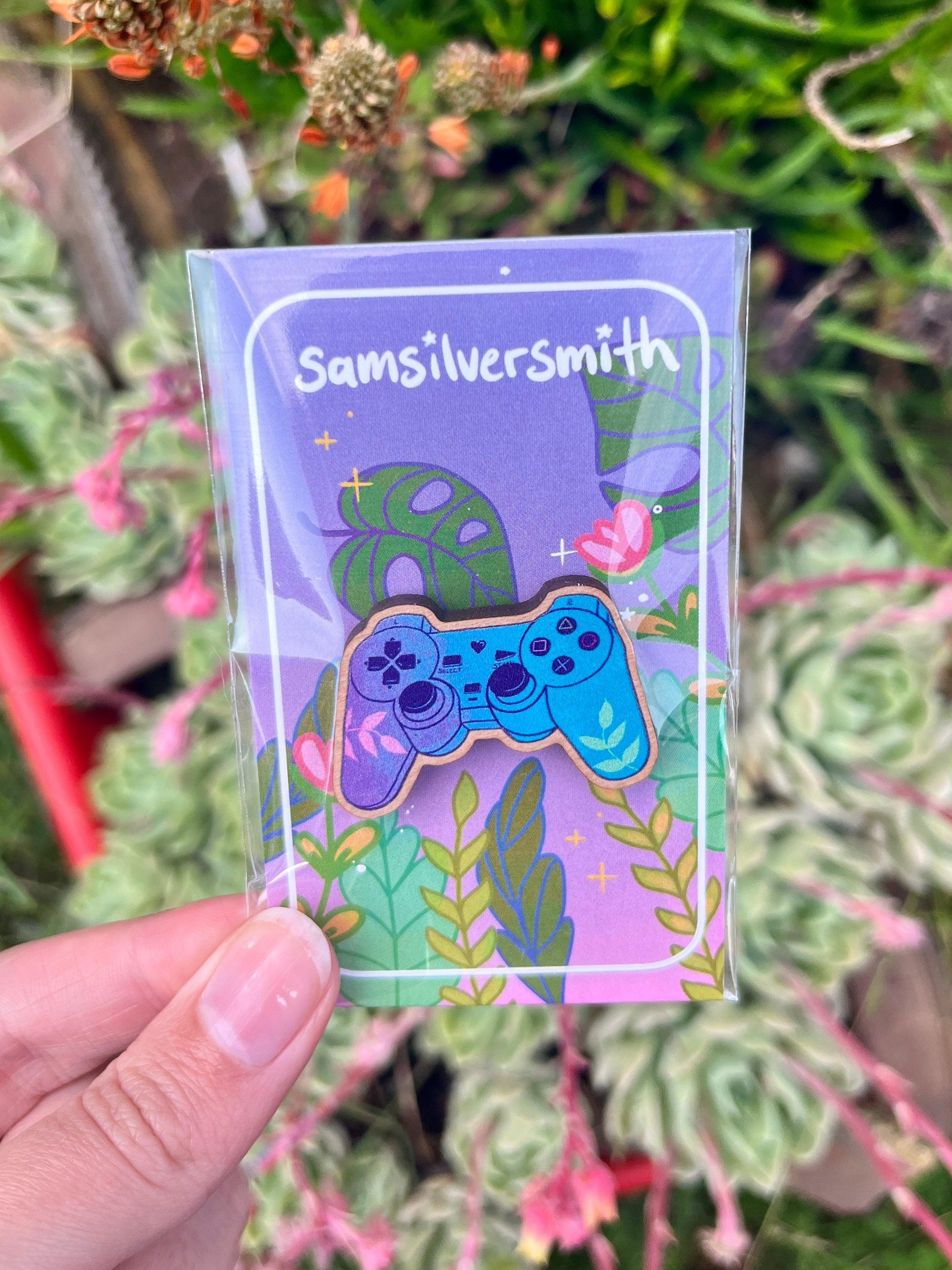 A hand holds a clear packet containing the artist-designed Playstation Controller Wooden Pin, featuring a colorful enamel design with a blue game controller set against vibrant leaves and abstract shapes. The packet, labeled Samsilversmith, beautifully showcases this charming piece.