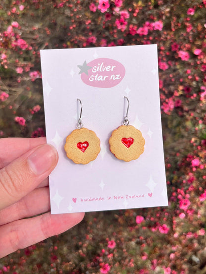 A hand displays a card featuring the Shrewsbury Polymer Clay Earrings, expertly crafted to resemble cookies adorned with red hearts. The card is inscribed with "silver star.nz" and highlights the distinct craftsmanship from New Zealand, while gentle pink flowers in the background enhance the scene's allure.