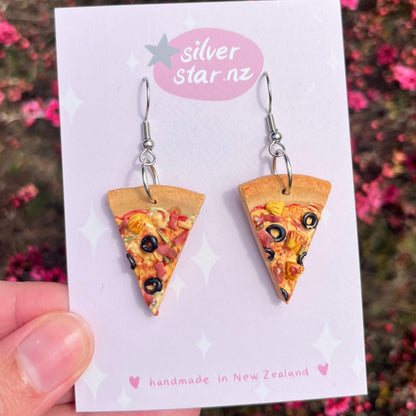 A hand displays a card featuring the Pizza Polymer Clay Earrings, designed with toppings such as olives and pineapple. The card is branded with the logo "silver star.nz," emphasizing this handcrafted New Zealand piece with stainless steel hooks, all set against a softly blurred floral backdrop.