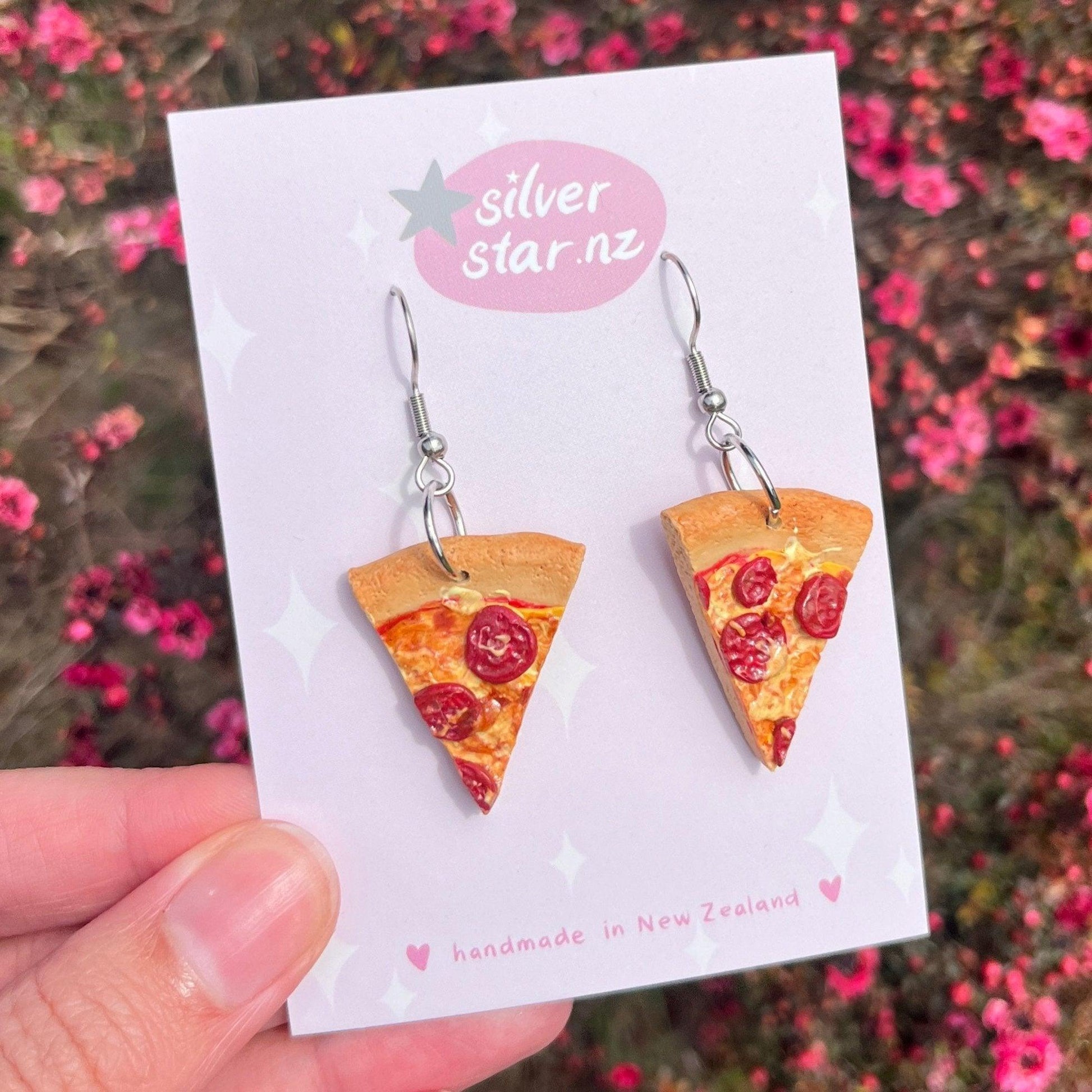 A close-up showcases a hand holding a white card featuring the Pizza Polymer Clay Earrings, intricately designed with pepperoni and cheese details. The card is adorned with a star and reads "silver star.nz" alongside "handmade in New Zealand." In the background, pink flowers enhance the charm of this playful design.