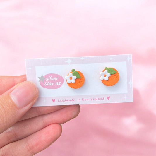 In a soft pink backdrop, a hand displays the Orange Blossom Polymer Clay Stud Earrings, designed in the shape of small round oranges topped with delicate white flowers. These handmade earrings from New Zealand come attached to a card labeled "silver star.nz" and "handmade in New Zealand.