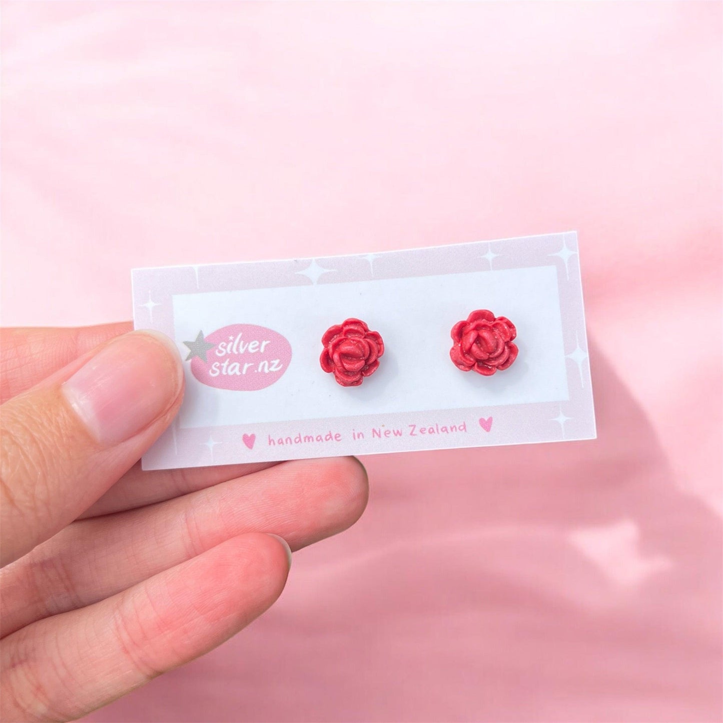 A hand holds a card displaying a pair of Rose Polymer Clay Stud Earrings, featuring two red flower-shaped earrings with stainless steel hooks. The card showcases a star design alongside the text "silver star nz" and "handmade in New Zealand." The soft, light pink fabric background accentuates the unique design of these handmade earrings.