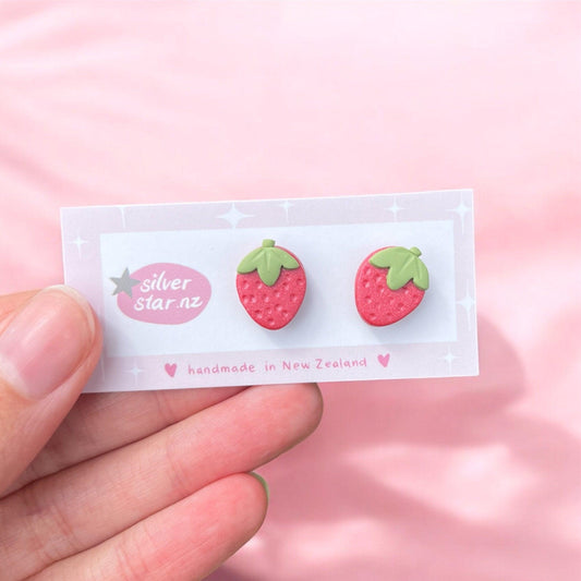 A person's hand holds a card showcasing Strawberry Polymer Clay Stud Earrings, uniquely shaped like strawberries with green leaves. The card prominently displays the silver star.nz logo and includes a note indicating that these products are handmade in New Zealand. A soft pink background lends a delicate touch to the scene.