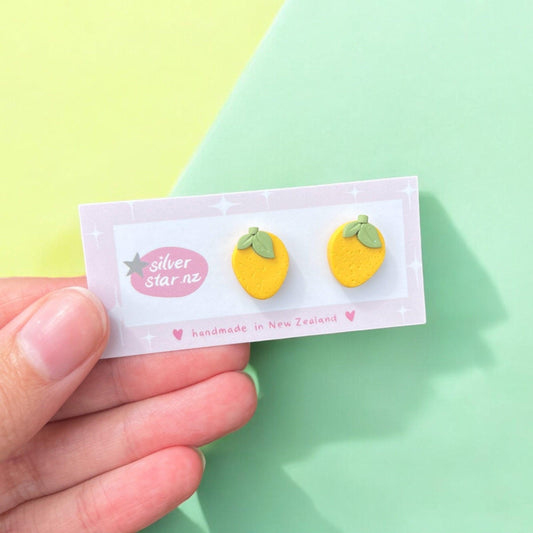 A hand displays a card featuring a pair of handmade lemon polymer clay stud earrings with green leaves. The card, embellished with decorative hearts and stars, reads "silver star z" and "handmade in New Zealand." Featuring stainless steel hooks, the background is a blend of light green and yellow.