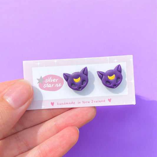 A close-up shot captures a hand presenting a card displaying two distinct purple cat-shaped earrings adorned with yellow crescent moons on their foreheads. The card reads "silver star.nz," highlighting the Luna Cat Polymer Clay Stud Earrings, which are handmade in New Zealand. The background is a soft shade of purple.