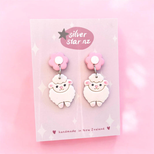 Displayed on a pink card, the Sheep Polymer Clay Earrings showcase adorable cartoon-style sheep with pink flower studs. The card is inscribed with "silver star.nz" and "handmade in New Zealand," set against a soft pink backdrop adorned with star patterns. Enjoy this unique design!