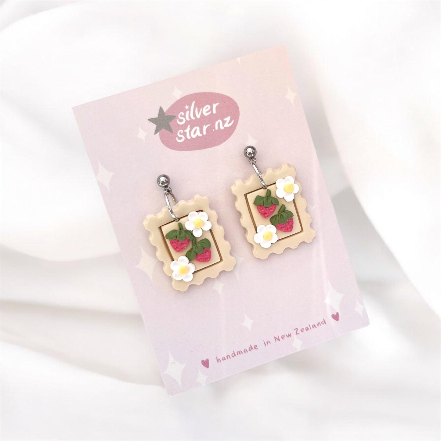 Introducing the Strawberry Stamp Polymer Clay Earrings, uniquely designed with postage stamp shapes featuring vibrant strawberries and daisies. These beautiful handmade earrings hang on a pink card labeled silver star.nz and are crafted in New Zealand with durable stainless steel hooks.