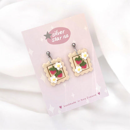 Introducing the Strawberry Stamp Polymer Clay Earrings, uniquely designed with postage stamp shapes featuring vibrant strawberries and daisies. These beautiful handmade earrings hang on a pink card labeled silver star.nz and are crafted in New Zealand with durable stainless steel hooks.