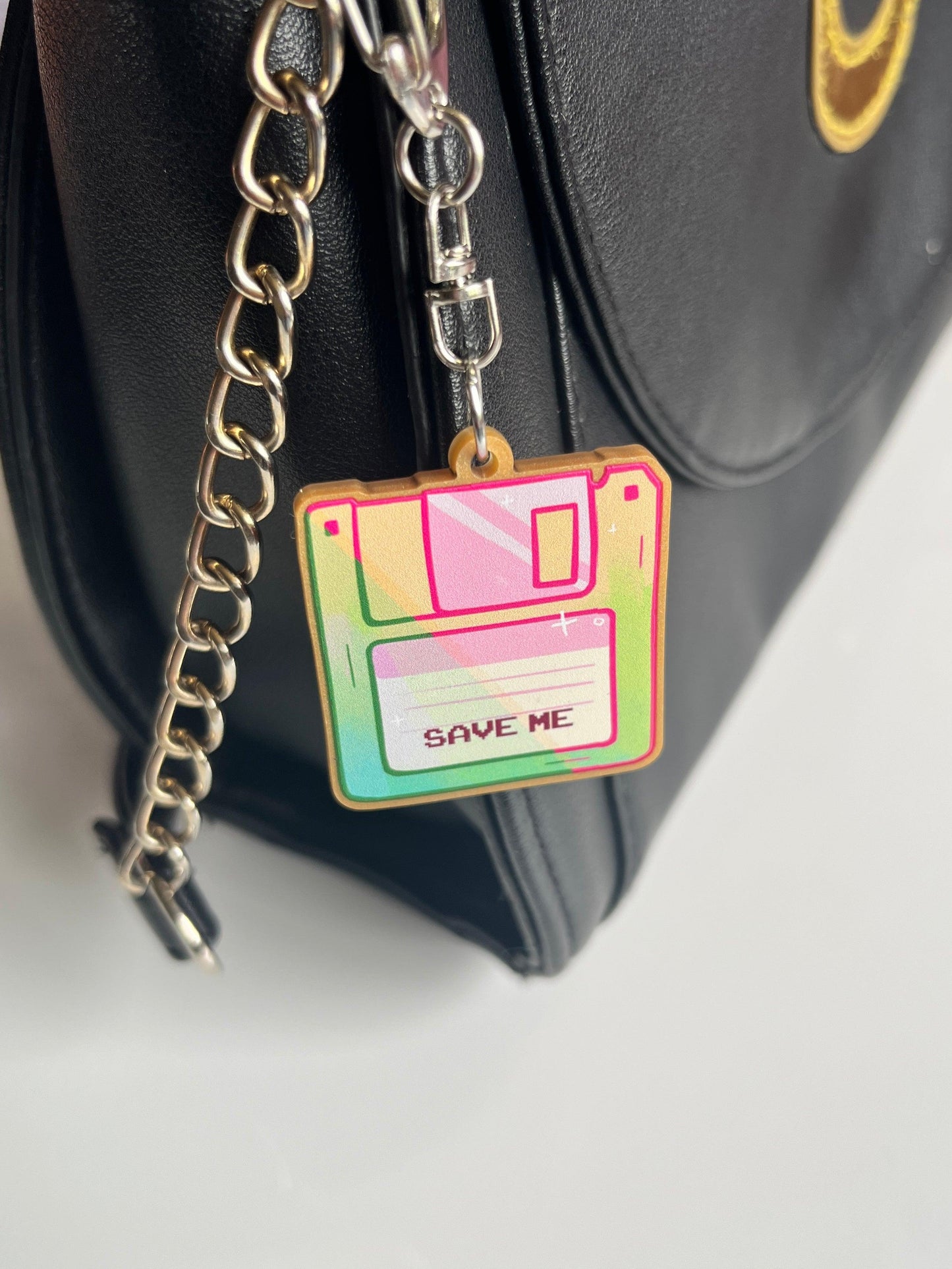 Close-up of a black bag with a chain strap, showcasing the Floppy Disk Acrylic Keyring, artistically designed in vibrant colors and labeled with "SAVE ME" at the center.