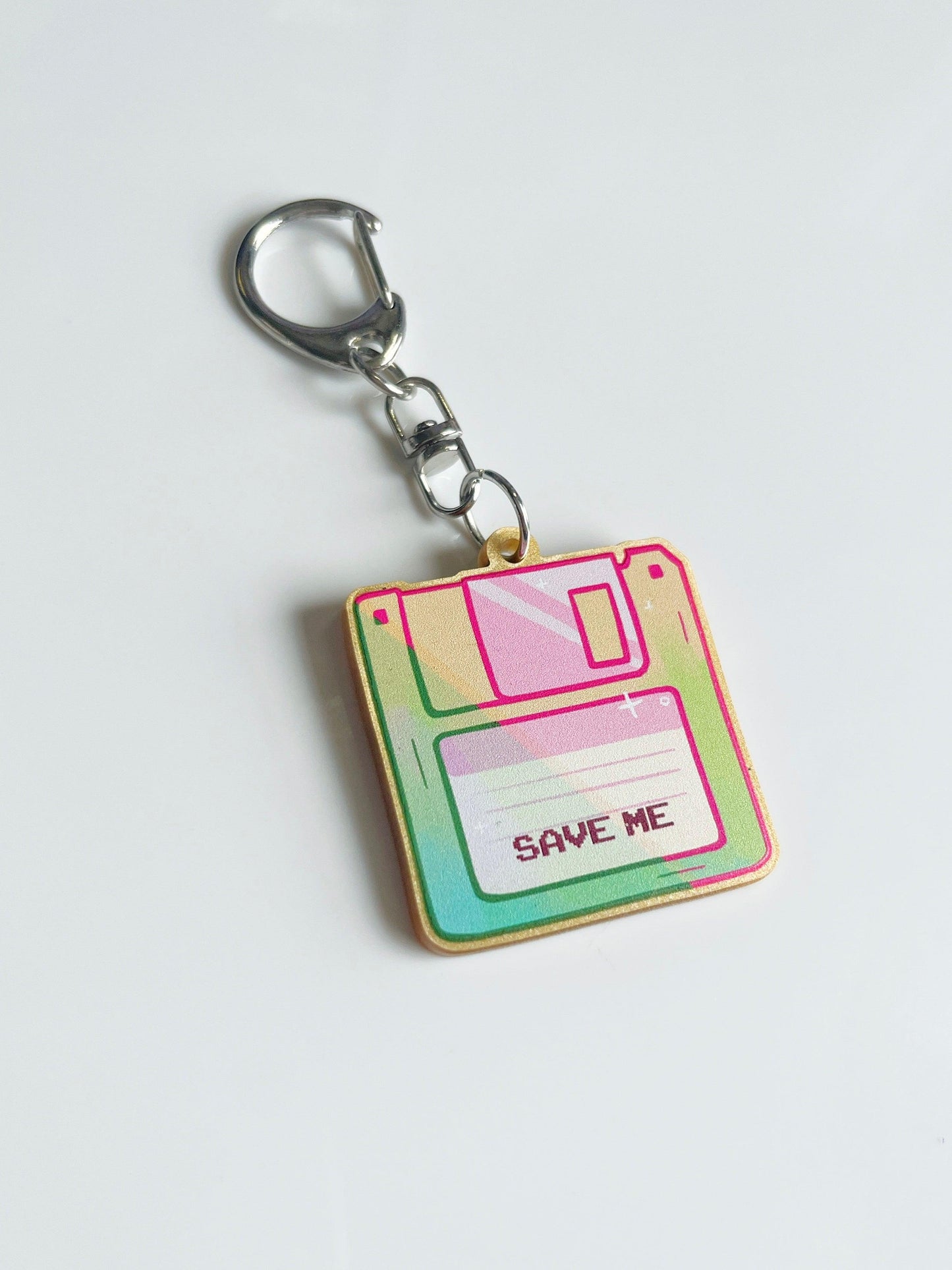 The Floppy Disk Acrylic Keyring, artist-designed with the vibrant phrase "SAVE ME," is elegantly crafted from acrylic and attached to a sleek silver metal ring. It rests stylishly on a white surface.