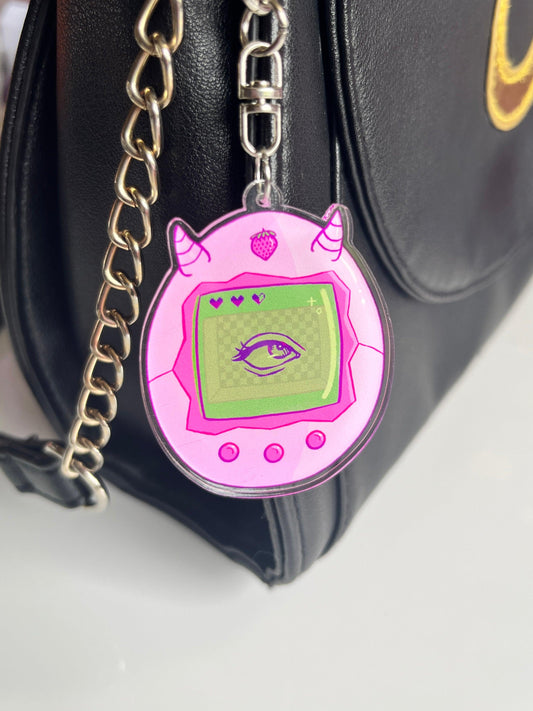 A chain on a black bag holds a Strawberry Tamagotchi Acrylic Keyring, featuring an artist-designed strawberry with small horns. The device's screen displays an eye against a pixelated green background, accompanied by three hovering hearts.