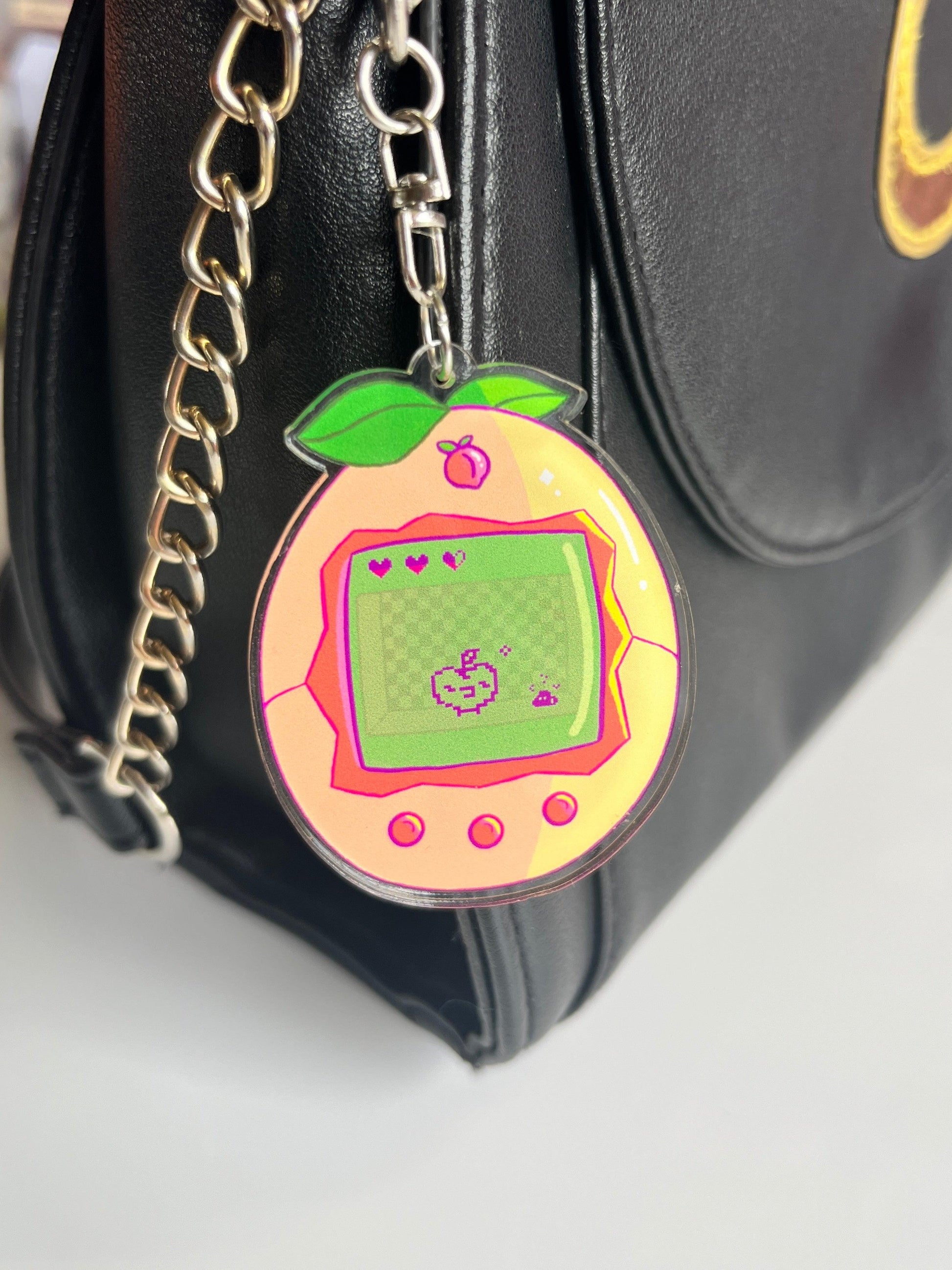 The Peach Tamagotchi Acrylic Keyring, featuring retro pixel art in the shape of a peach, hangs from a silver chain attached to a black leather bag.