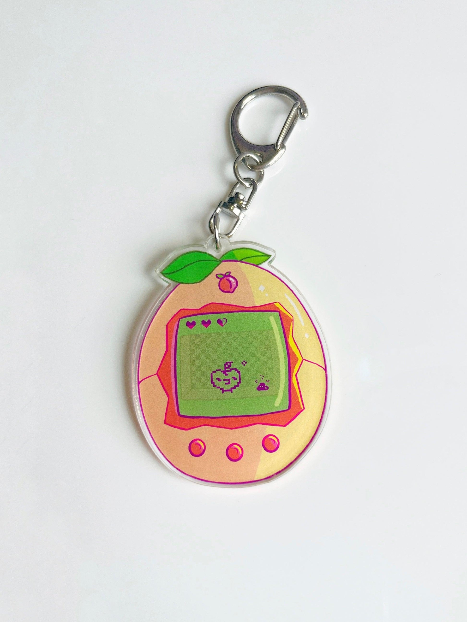 The Peach Tamagotchi Acrylic Keyring features a delightful digital pet device design, with a small screen displaying pixel art above three pink buttons. The metal loop at the top makes it easy to attach to various items.