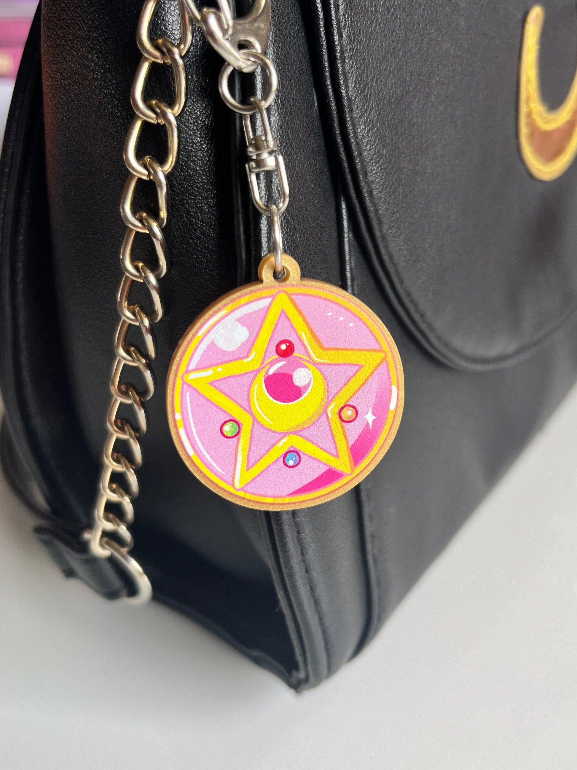 A black bag with a gold chain strap features the artist-designed Sailor Moon Locket Acrylic Keyring. This colorful pink and yellow keychain showcases a circular star design with a crescent moon and small decorative embellishments, adding a unique touch to your accessory collection.