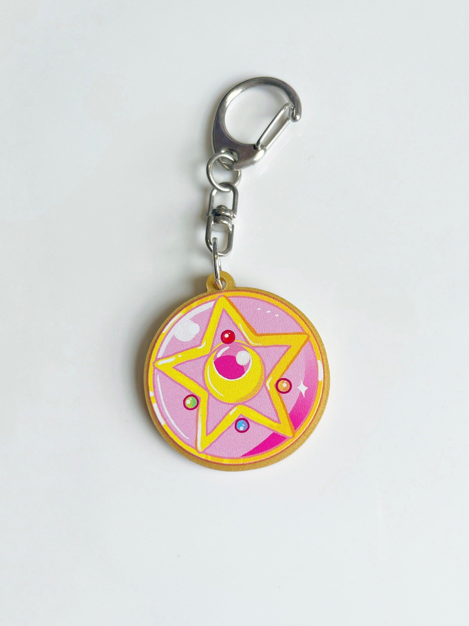 Introducing the Sailor Moon Locket Acrylic Keyring: an artist-designed creation featuring a pink circular charm adorned with a yellow star and colorful jewels. Inspired by a magical theme, this enchanting piece merges whimsical aesthetics with functionality, set against a clean white backdrop.