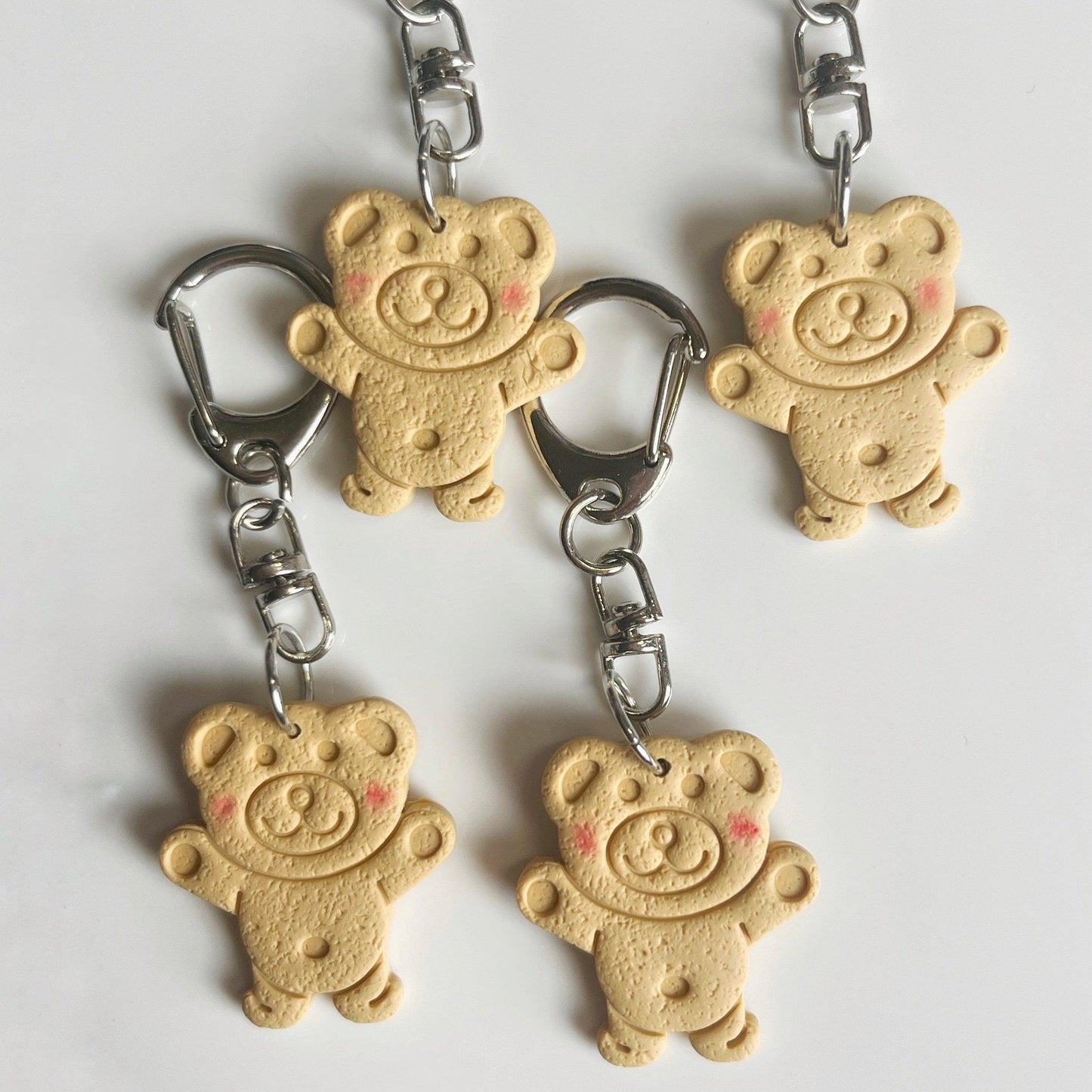 A collection of five Tiny Teddy Polymer Clay Keyrings, featuring a cheerful expression and biscuit-like textured surface, are arranged on a white background. These handmade keychains from New Zealand have a beige teddy bear shape with rosy cheeks and metal clasp attachments, making them charming gifts.