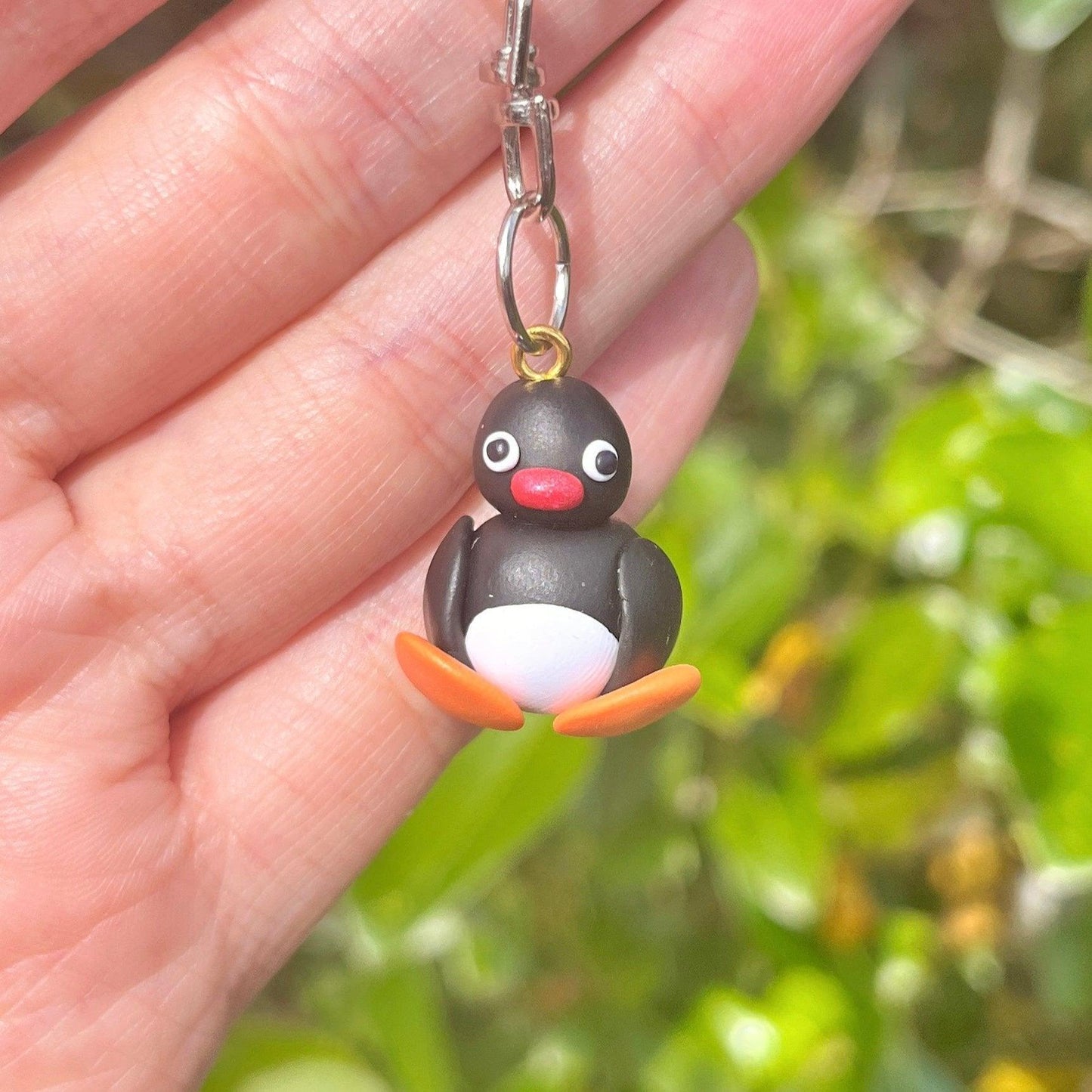 A person is holding a Pingu Polymer Clay Keyring, featuring a charm shaped like a cartoon penguin with a red beak and orange feet, crafted from durable lightweight material, set against a blurred green leafy background.