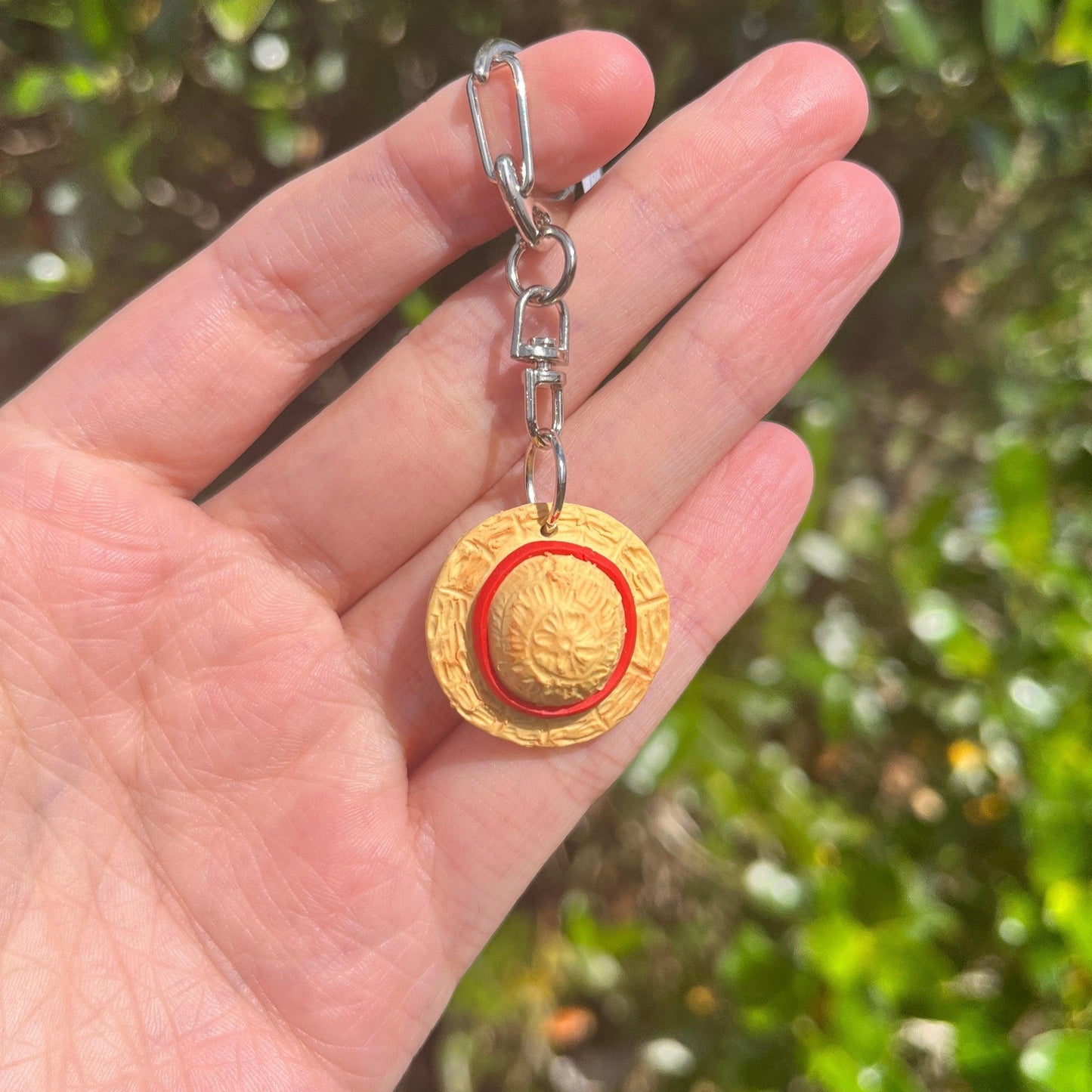 A hand holds the Luffy Hat One Piece Polymer Clay Keyring, showcasing its unique and funky design with a round, embossed medallion featuring a sun relief and red circle. Crafted from durable materials, this handmade polymer clay keyring stands out against the blurred greenery in the background.