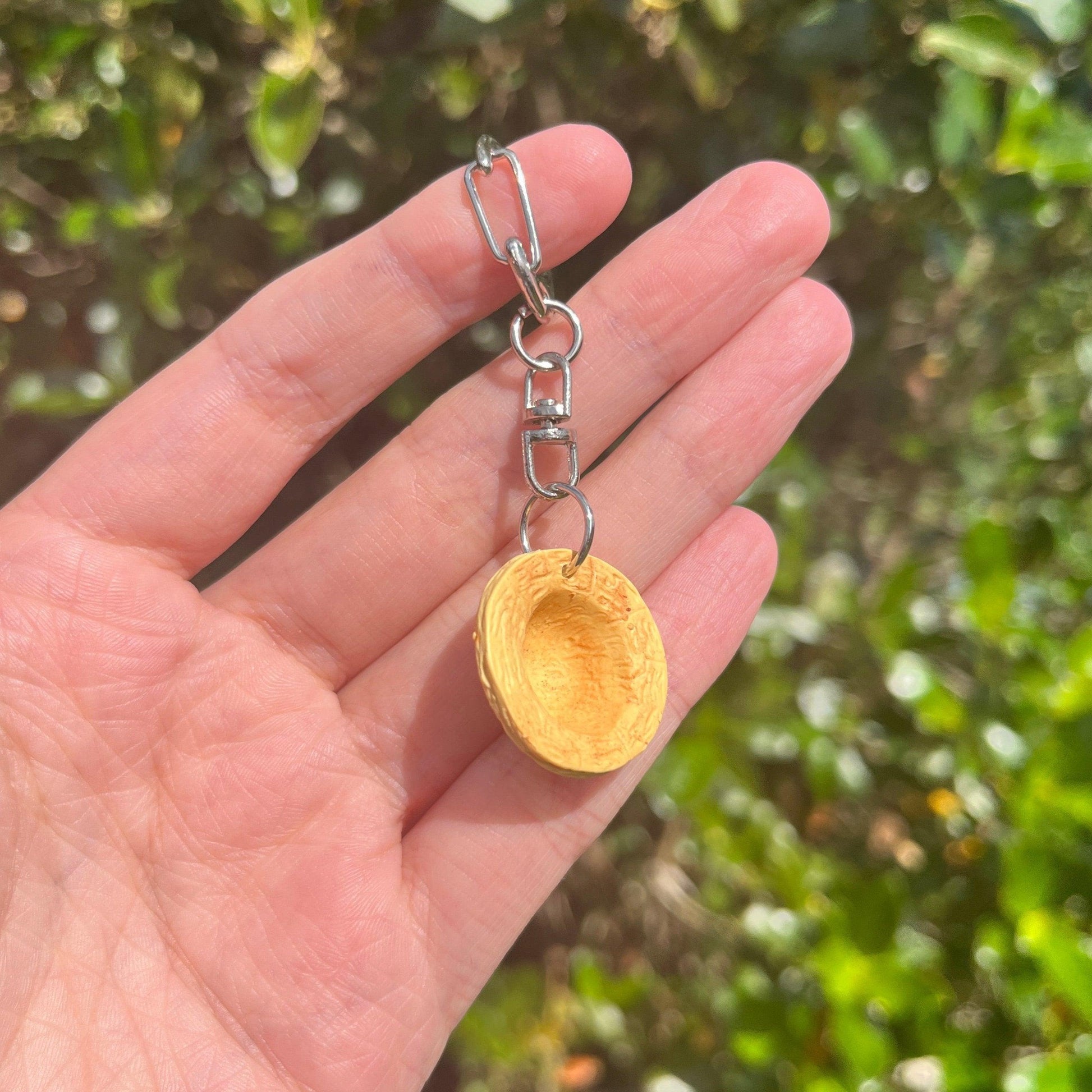 A hand holding the Luffy Hat One Piece Polymer Clay Keyring, featuring a small, round wooden charm in a unique funky design. The blurred green foliage in the background enhances the allure of these handmade keyrings.