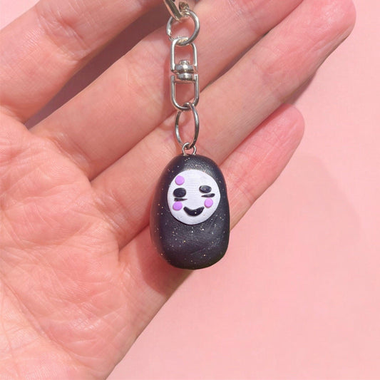 A hand holds the No Face Polymer Clay Keyring, showcasing a small, black charm shaped like a character with a smiling white face and purple spots on its cheeks. This charming piece stands out beautifully against the light pink background.