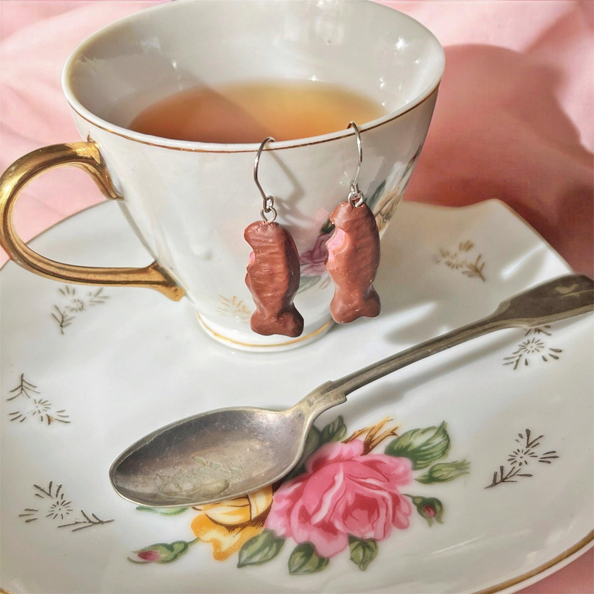 Handmade Mini Chocolate Fish Polymer Clay Earrings dangle from a teacup filled with tea. The teacup and saucer, adorned with floral designs, hold a silver teaspoon. The unique design stands out against a backdrop of pink fabric, creating a charming visual.