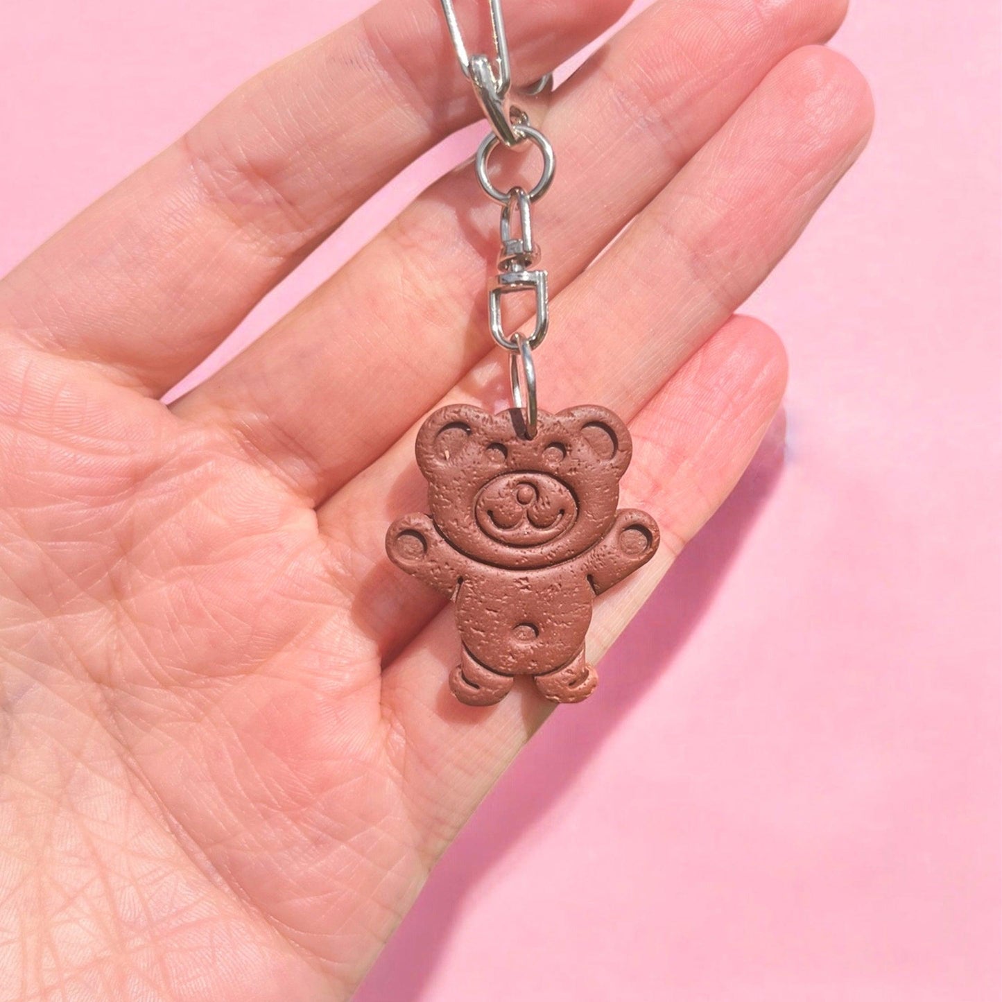 A hand displays the distinctive Tiny Teddy Polymer Clay Keyring, showcasing a small teddy bear with a textured finish and a cheerful smile against a pink background. This keyring, crafted as part of New Zealand handmade gifts, features a metal clasp attached to the bear's head.