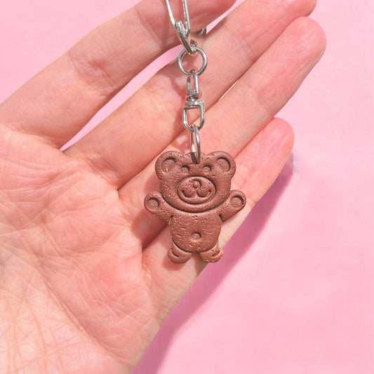 A hand displays the distinctive Tiny Teddy Polymer Clay Keyring, showcasing a small teddy bear with a textured finish and a cheerful smile against a pink background. This keyring, crafted as part of New Zealand handmade gifts, features a metal clasp attached to the bear's head.