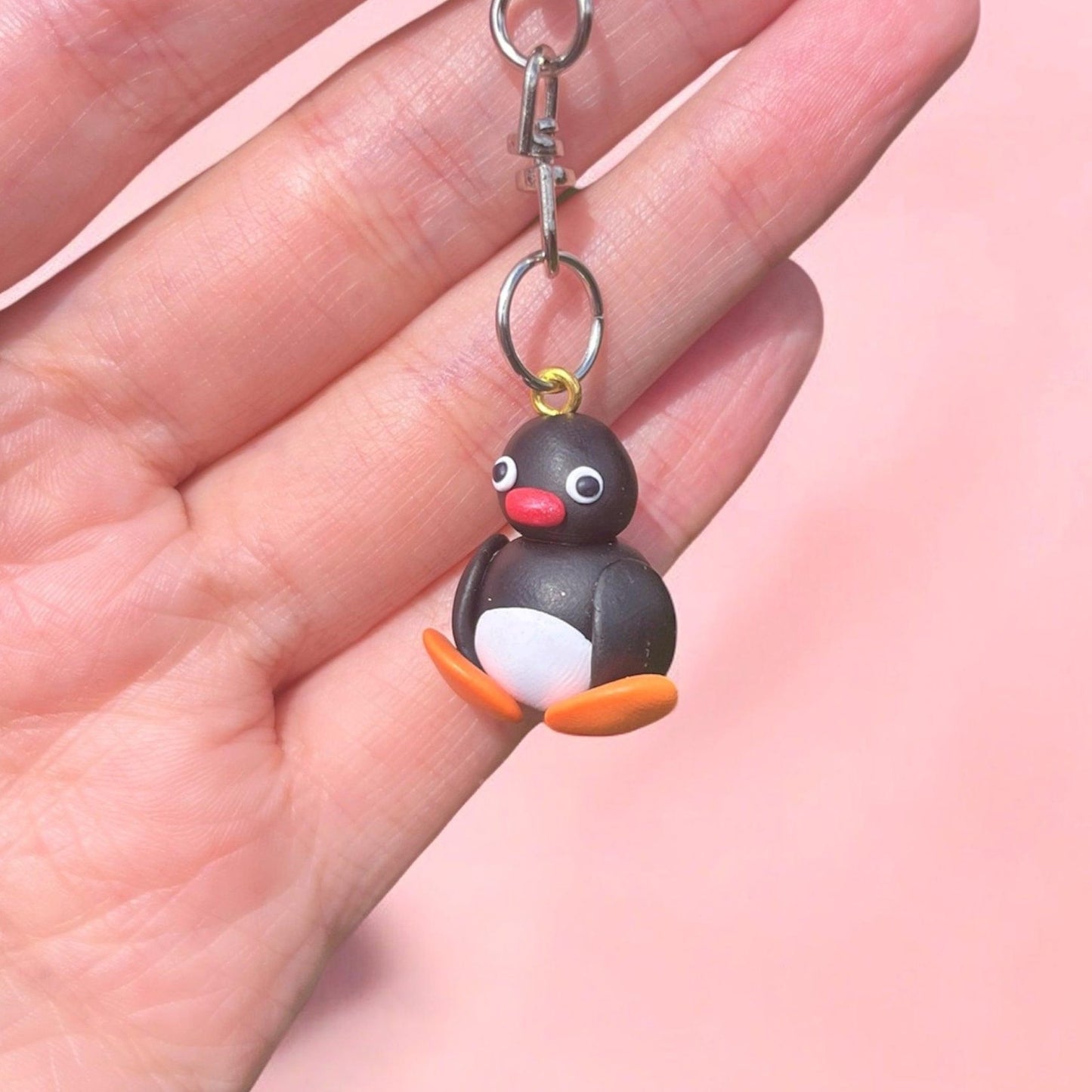 A hand holds the Pingu Polymer Clay Keyring featuring a small penguin figure crafted from durable lightweight material, showcasing wide eyes, a red beak, white belly, and orange feet against a soft pink background.