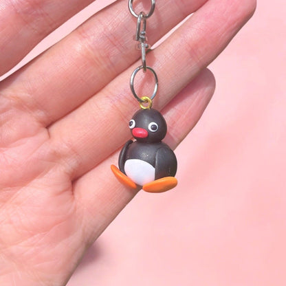 A hand holds the Pingu Polymer Clay Keyring featuring a small penguin figure crafted from durable lightweight material, showcasing wide eyes, a red beak, white belly, and orange feet against a soft pink background.