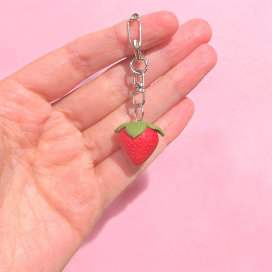 A hand holds the Strawberry Polymer Clay Keyring by silverstar.nz, showcasing a unique design with a strawberry-shaped charm against a soft pink background. The charm, crafted from handmade polymer clay, features a red strawberry with green leaves, complemented by a silver keychain—ideal as a perfect gift for a loved one.