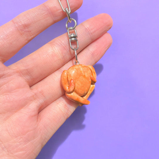 A hand is holding the Roast Chicken Polymer Clay Keyring, which is shaped like a roasted turkey and crafted from durable material, against a solid purple background. This handmade keychain features a metal clasp for easy attachment, showcasing its unique funky design.