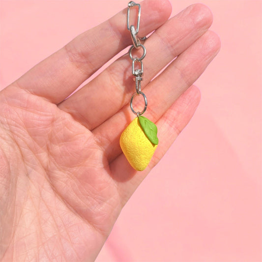 A hand holding the Lemon Polymer Clay Keyring, which features a small lemon charm crafted from durable and lightweight material with a green leaf against a soft pink background.