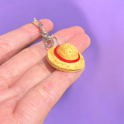 The Luffy Hat One Piece Polymer Clay Keyring, featuring a small figurine of a straw hat with a red band crafted from durable material, is held between fingers. The solid purple background highlights its unique funky design.