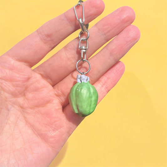 Against a bright yellow background, a hand displays the Feijoa Polymer Clay Keyring, which features a small green fruit crafted with a unique funky design from handmade polymer clay.