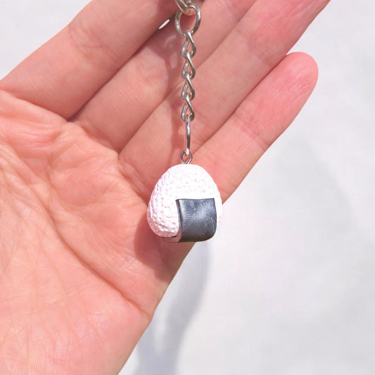 An Onigiri Polymer Clay Keyring, made from durable material and featuring a textured surface with a black seaweed strip, rests in an open palm. Its unique design stands out against the soft, blurred white background.