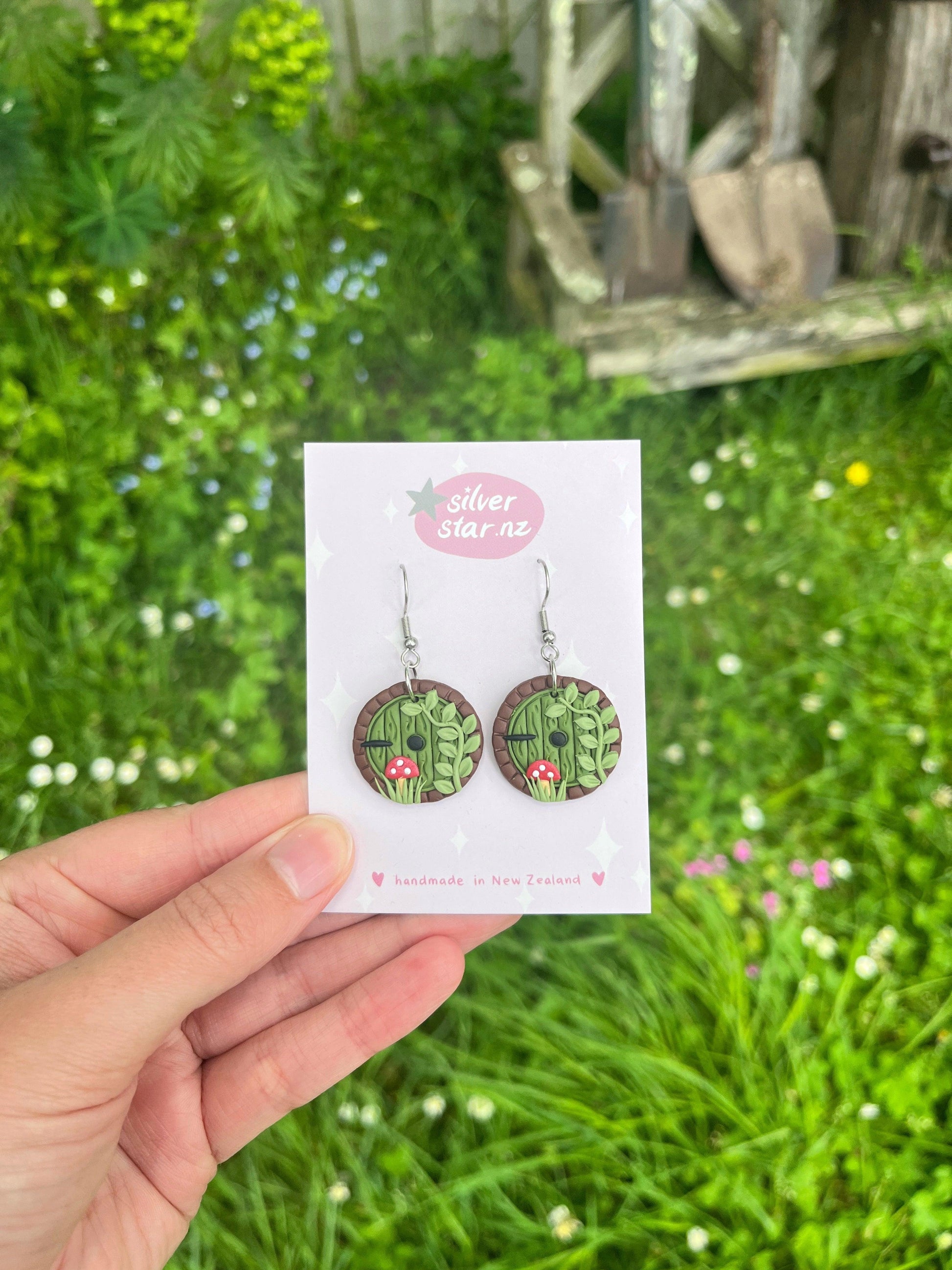 A hand holds up a card displaying the "Hobbit Door Polymer Clay Earrings," adorned with green leaf motifs and small red mushrooms. These handmade earrings elegantly dangle from stainless steel hooks, set against a lush grass background dotted with white flowers. The card reads "silver star nz.