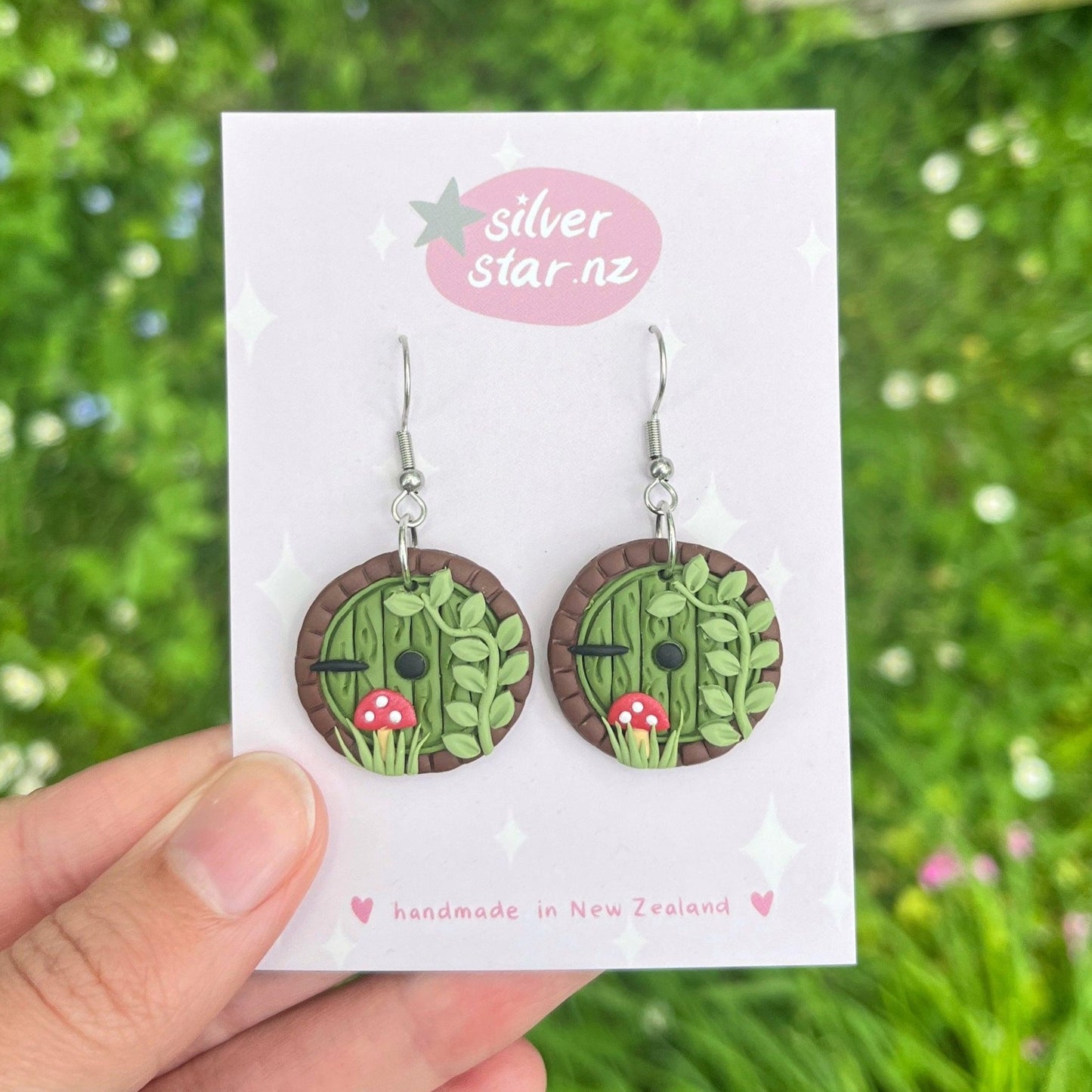 A hand presents a pair of round, handmade Hobbit Door Polymer Clay Earrings on a card with silver star.nz branding. These earrings feature a small green door framed by plants and mushrooms, crafted from polymer clay. With stainless steel hooks, they are set against a backdrop of blurred greenery.