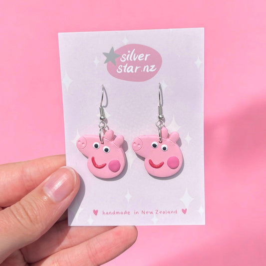 A hand holds the Peppa Pig Polymer Clay Earrings, showcasing pink cartoon pig faces with smiling expressions, crafted from handmade polymer clay and attached to a card labeled silver star.nz. Featuring stainless steel hooks, they stand out against the solid pink background.