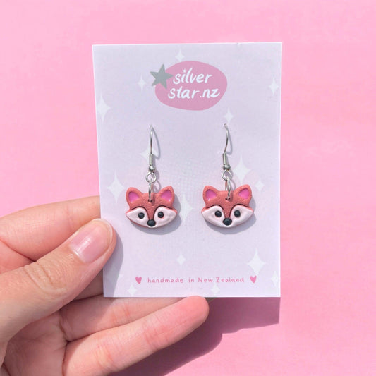 A hand displays a card against a lively pink background, featuring unique earrings designed as pink foxes. The card is labeled with "silver star.nz" accompanied by small star illustrations, emphasizing the Fox Polymer Clay Earrings handmade in New Zealand with stainless steel hooks.