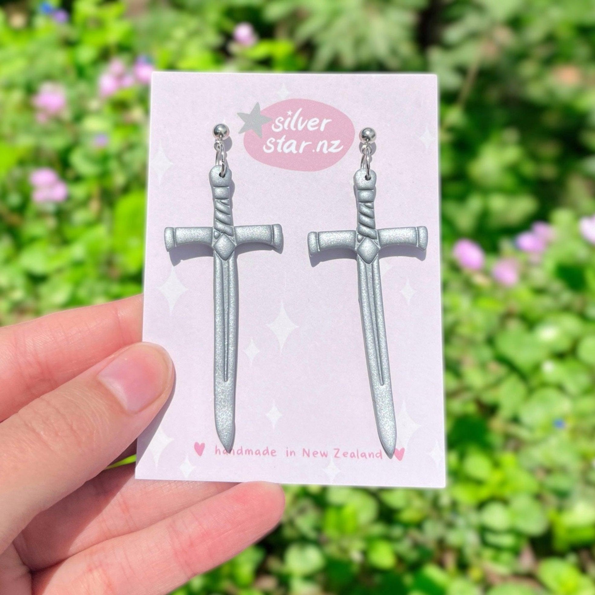 A hand displays a card showcasing the Long Silver Sword Polymer Clay Earrings. The card, labeled with "silver star.nz," emphasizes that these earrings are handmade in New Zealand. The unique design is accentuated by a backdrop of green foliage and pink flowers.