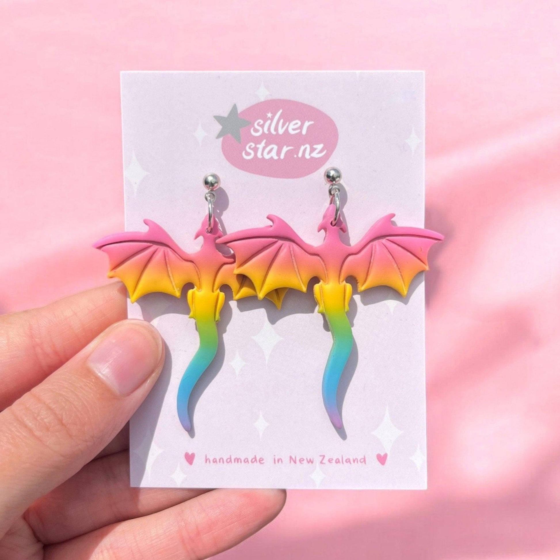 A hand holds a card featuring the Rainbow Dragon Polymer Clay Earrings, showcasing a unique design with pink, yellow, and blue gradients. The card is labeled Silver Star.nz and highlights these handmade earrings from New Zealand against a pink background.