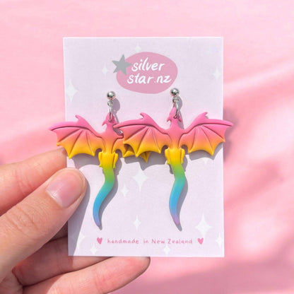 A hand holds a card featuring the Rainbow Dragon Polymer Clay Earrings, showcasing a unique design with pink, yellow, and blue gradients. The card is labeled Silver Star.nz and highlights these handmade earrings from New Zealand against a pink background.