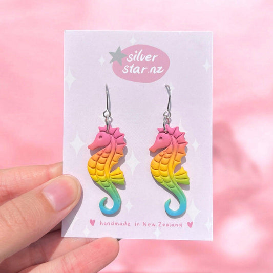 A hand presents a card featuring the Rainbow Seahorse Polymer Clay Earrings in vibrant hues of pink, orange, yellow, green, and blue. Labeled with "silver star.nz" against a pink backdrop, these handmade earrings showcase an exquisite design crafted in New Zealand.