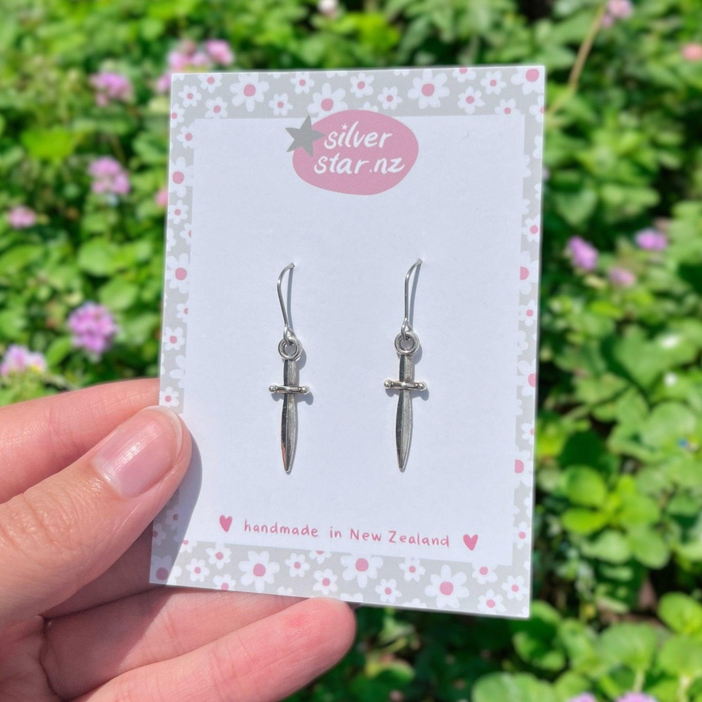 A hand holds a card displaying the Mini Silver Dagger Earrings from silver star.nz, featuring stainless steel hooks. The card showcases a floral border and "handmade in New Zealand" text. A blurred green and pink floral background complements the lightweight earrings' charm.