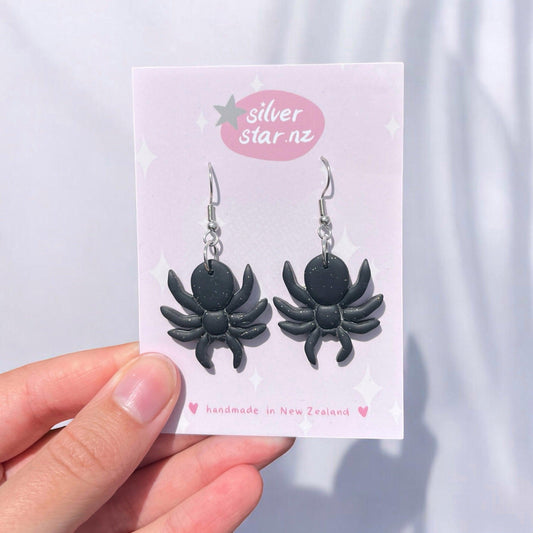 A hand showcases a pair of funky, Spider Polymer Clay Earrings, crafted in black spider shapes. They are displayed on a pink card featuring "silverstar.nz" and emphasizing their handmade origin in New Zealand, all set against a softly lit background.